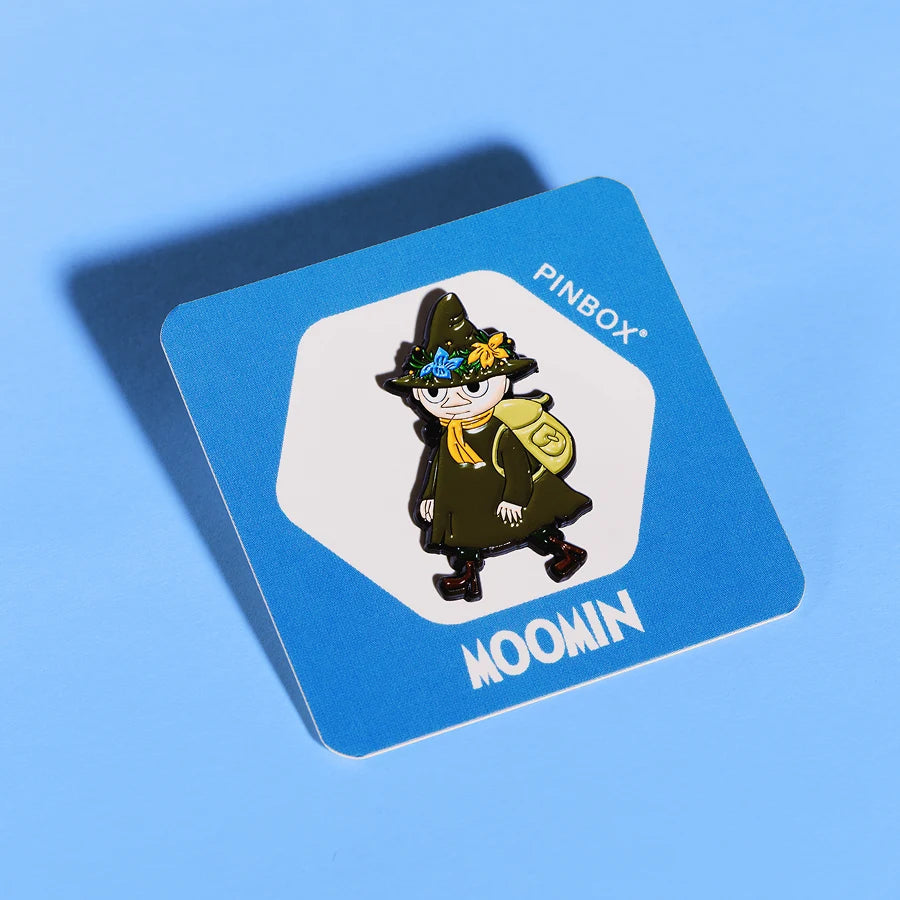 Emaliowana pin przedstawiająca Włóczykija w zielonym stroju podróżnika z kapeluszem ozdobionym kwiatami, żółtym szalikiem i zielonym plecakiem. Przypięta do niebieskiej karteczki z napisem Moomin